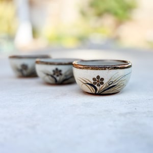 May include: Three small, off-white ceramic bowls with a floral design. Each bowl features a dark brown flower with black and tan accents. The rims are trimmed with a gold-coloured band. The bowls are arranged in a row, with one in focus.