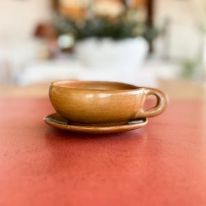 Może przedstawiać: Ręcznie wykonany, brązowy zestaw filiżanki i spodka ceramicznego. Filiżanka ma zaokrąglony kształt z uchwytem i stoi na pasującym spodku. Zestaw umieszczony jest na czerwonej powierzchni z rozmytym tłem.