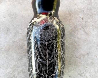 May include: A black ceramic vase with a glossy finish. The vase features a detailed white floral and leaf pattern etched into the surface. The vase is approximately 8 inches tall and has a narrow neck.