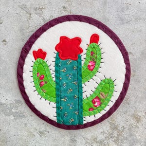 May include: Round, quilted cactus coaster with a white background and a dark purple border. The cactus is green with floral accents and a red flower. The coaster is handmade and features decorative stitching.