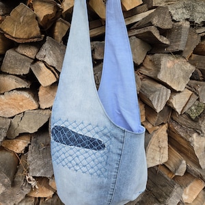Könnte beinhalten: Eine Jeans-Umhängetasche mit hellblauem Innenfutter und Trägern. Die Tasche hat einen Aufnäher mit einem genähten Netzdesign in einem dunkleren Blauton. Die Tasche befindet sich vor einem Hintergrund aus gestapeltem Brennholz.