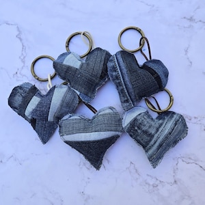 May include: A collection of six denim heart-shaped keychains. Each keychain is made from repurposed blue jean fabric, with varying shades of blue and gray. The keychains have bronze-colored metal rings and brown leather or fabric loops.