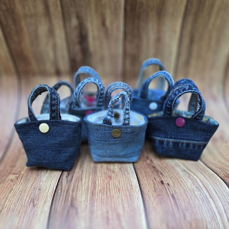 May include: A collection of small, miniature tote bags crafted from blue denim fabric. Each bag features two sturdy handles and a single snap button closure in various colors, including gold, bronze, and purple, secured on the front exterior.
