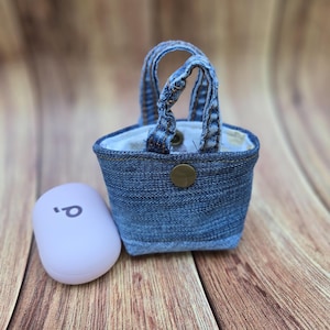 May include: A miniature denim tote bag with two handles, featuring a brass-colored metal snap button closure on the front. Beside the bag sits a small, smooth, light lavender-colored oval case with a dark logo printed on its top surface.