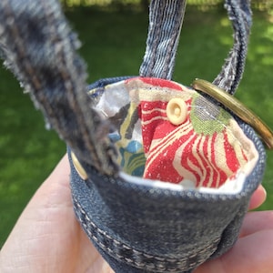 May include: A small pouch crafted from dark blue denim fabric with frayed edges and a handle. The interior is lined with a multi-colored floral fabric featuring red, white, and blue patterns. A circular brass ring is attached to the side.
