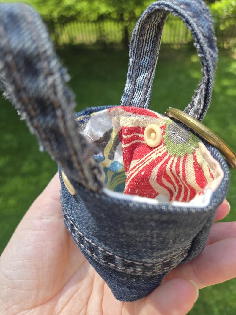 May include: A small pouch crafted from dark blue denim fabric with frayed edges and a handle. The interior is lined with a multi-colored floral fabric featuring red, white, and blue patterns. A circular brass ring is attached to the side.