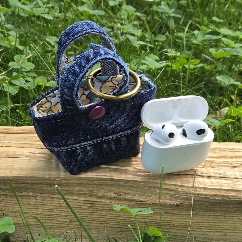 May include: A miniature dark blue denim tote bag with two handles, a purple snap button, and a floral fabric lining, featuring a gold-toned metal ring attached to the top. Next to it sits a white wireless earbud charging case with two earbuds.