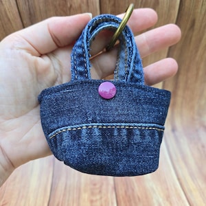 May include: A miniature denim handbag keychain featuring dark blue fabric with visible orange stitching, a single purple snap button closure, and a gold-toned metal ring attached to the top handle for secure fastening to keys or bags.