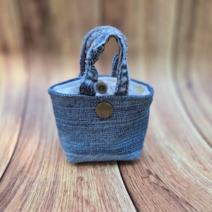 May include: A small, structured pouch crafted from blue denim fabric, featuring two short top handles made of matching denim. The exterior includes a single centered brass-colored metal snap button closure, while the interior is lined with white fabric.