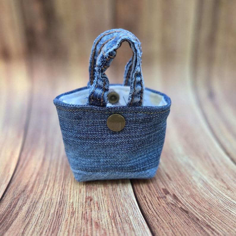 May include: A small, structured pouch crafted from blue denim fabric, featuring two short top handles made of matching denim. The exterior includes a single centered brass-colored metal snap button closure, while the interior is lined with white fabric.