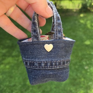 May include: A miniature tote bag crafted from dark blue denim fabric, featuring two frayed denim handles, a horizontal stitched seam detail across the front, and a small, smooth, cream-colored heart-shaped embellishment centered near the top edge.