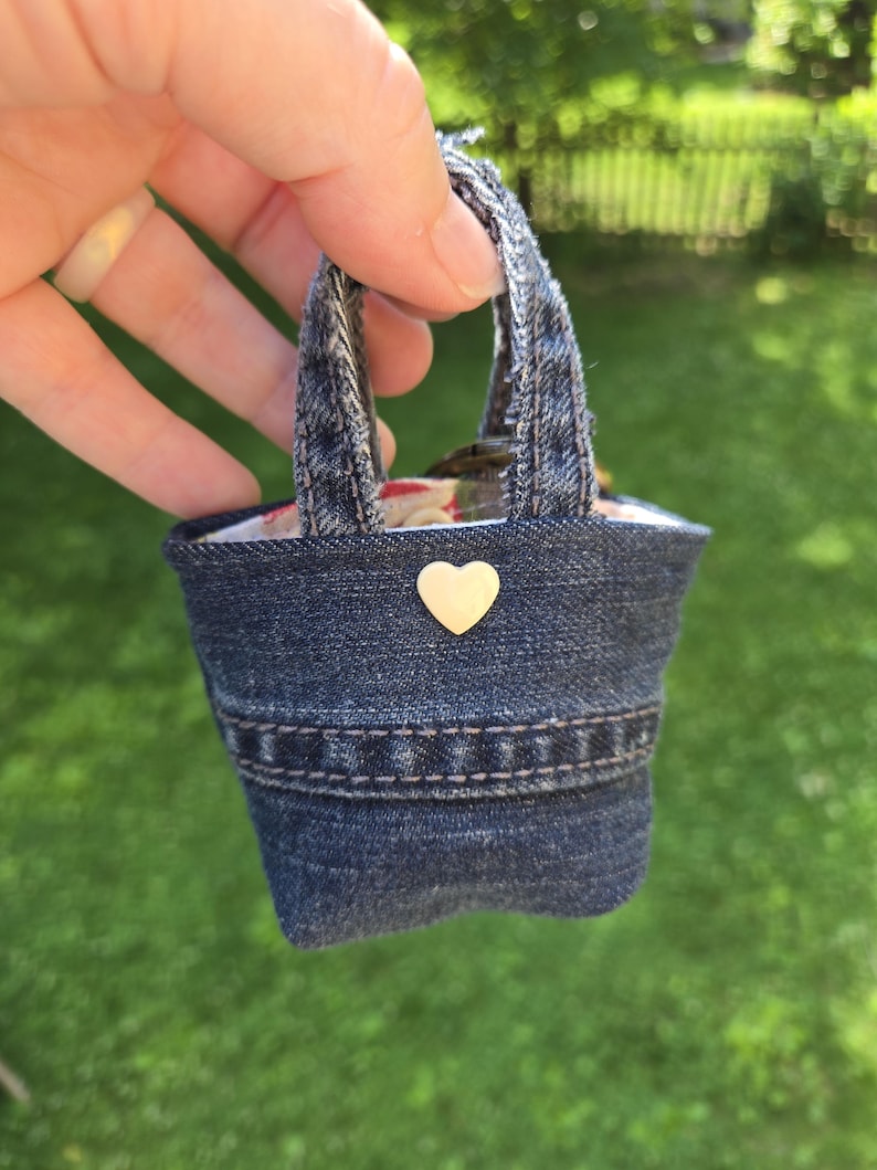 May include: A miniature tote bag crafted from dark blue denim fabric, featuring two frayed denim handles, a horizontal stitched seam detail across the front, and a small, smooth, cream-colored heart-shaped embellishment centered near the top edge.
