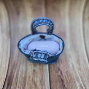 May include: A small, blue denim pouch with a top handle and a frayed fabric tab closure. The pouch features a light-colored interior lining and contains a smooth, light pink, oval-shaped electronic charging case nestled inside the main compartment.