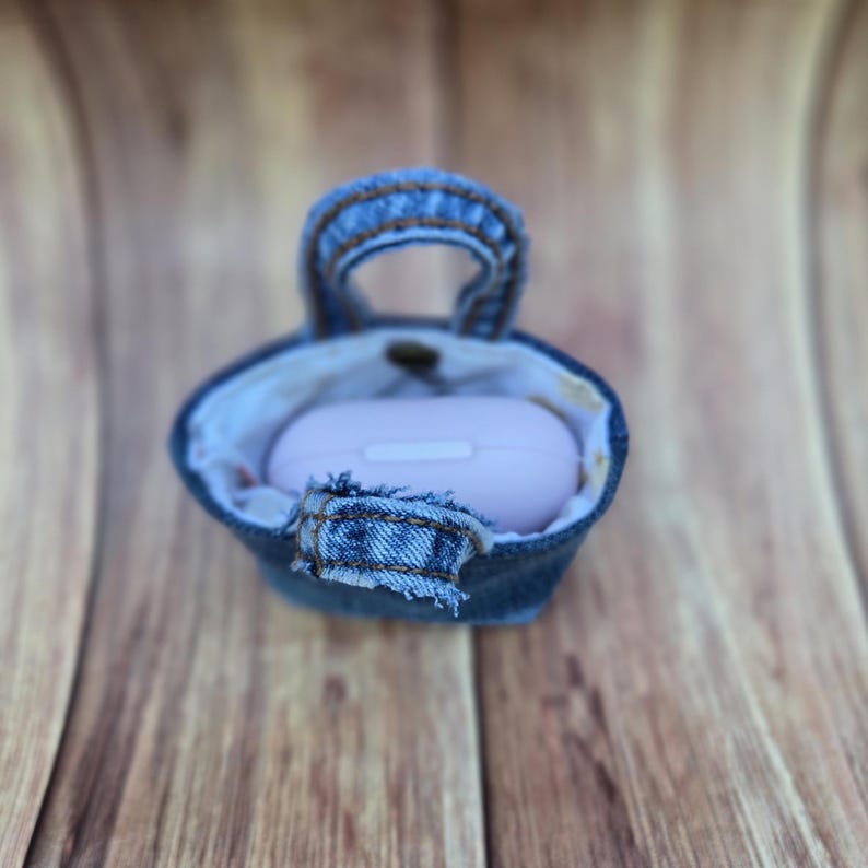 May include: A small, blue denim pouch with a top handle and a frayed fabric tab closure. The pouch features a light-colored interior lining and contains a smooth, light pink, oval-shaped electronic charging case nestled inside the main compartment.