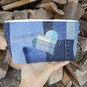 May include: A rectangular zippered pouch constructed from various shades of blue denim fabric in a patchwork design. The front features a central heart-shaped applique made of plaid fabric, accented with white hand-stitched embroidery details.