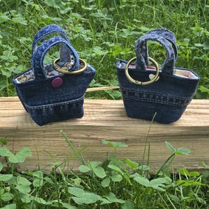 May include: Two miniature tote bags crafted from blue denim fabric. Each bag features two small handles, a brass-toned metal ring attachment, and a patterned fabric lining. One bag includes a small purple snap button closure on the front exterior.