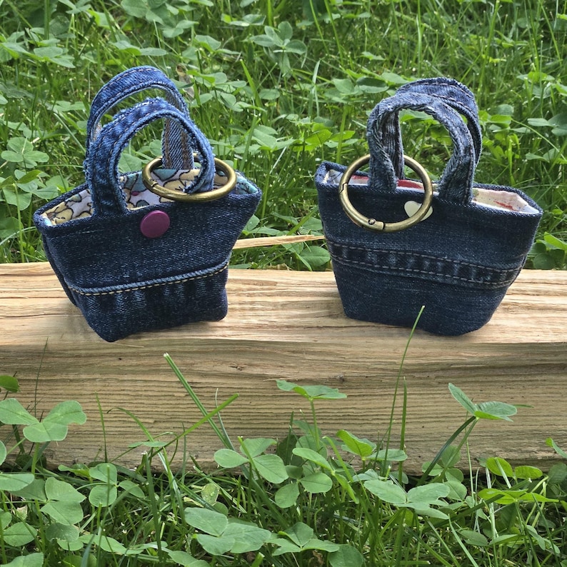May include: Two miniature tote bags crafted from blue denim fabric. Each bag features two small handles, a brass-toned metal ring attachment, and a patterned fabric lining. One bag includes a small purple snap button closure on the front exterior.