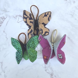 May include: Four handmade butterfly hair accessories in various colors and patterns. The butterflies feature brown, green, and purple wings with decorative stitching and elastic bands for attachment. Each butterfly has a beaded body.