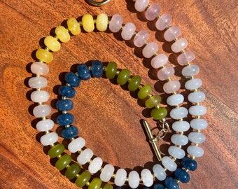 May include: A colorful beaded necklace with a gold clasp. The necklace features a gradient of colors, including yellow, pink, blue, and green beads. The beads are strung on a gold chain, creating a vibrant and eye-catching accessory.
