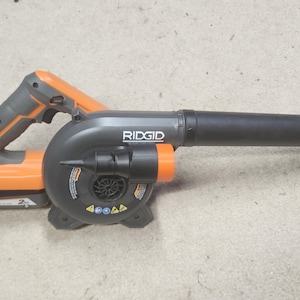 Könnte beinhalten: Ein schwarzer und orangefarbener RIDGID 18V Akku-Laubbläser mit schwarzer Düse. Der Gebläse hat einen Abzugsschalter und eine Batterieanzeige.