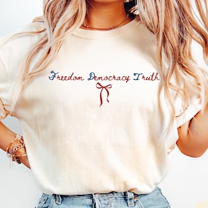 Op de afbeelding: Ivoor kleurig t-shirt met de woorden "Freedom Democracy Truth" in blauw en rood script, en een rode strik afbeelding. Het shirt is gemaakt van een zacht materiaal.