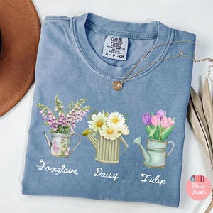 Può includere: Una maglietta blu con una grafica di tre annaffiatoi con fiori. Gli annaffiatoi sono etichettati "Foxglove", "Daisy" e "Tulip".