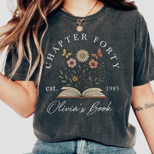 May include: Dark gray t-shirt with the text "CHAPTER FORTY" and "Olivia's Book" in white script. The shirt features a floral design with butterflies and an open book. The year "1985" is also printed on the shirt.