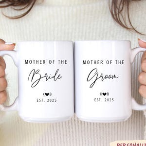 Puede incluir: Dos tazas de cerámica blanca con texto negro que dice "Mother of the Bride" y "Mother of the Groom". Cada taza tiene las iniciales "K♥B" y "EST. 2025". Las tazas son personalizadas.