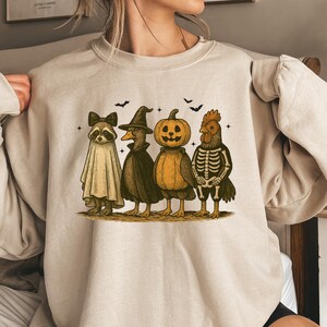 Puede incluir: Sudadera color arena con un diseño temático de Halloween. El diseño incluye cuatro personajes disfrazados: un mapache con disfraz de fantasma, un pato con disfraz de bruja, una calabaza y un esqueleto de pollo. También se incluyen murciélagos y estrellas.