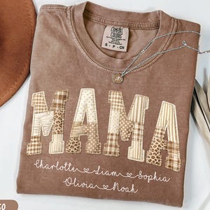 Puede incluir: Camiseta color espresso con la palabra "MAMA" en letras de patchwork. Debajo están bordados nombres. Un collar plateado con un colgante descansa sobre la camiseta. Un sombrero marrón y hojas secas están en el fondo.