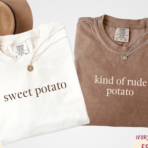 May include: Two t-shirts, one ivory and one espresso, with the phrases "sweet potato" and "kind of rude potato" printed on them. Each shirt has a gold necklace. A brown hat is visible in the background.