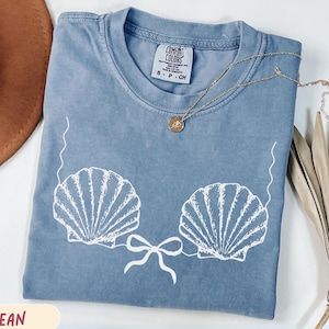 Op de afbeelding: Een blauw spijkerbroekkleurig t-shirt met een wit schelpmotief. Het shirt heeft een ronde hals en korte mouwen. Een gouden ketting met een hanger ligt op het shirt. Een bruine hoed en gedroogde bladeren zijn op de achtergrond.