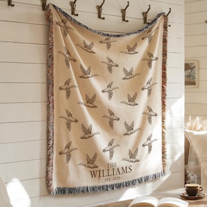 May include: A cream-colored woven throw blanket featuring a repeating pattern of dark brown flying ducks. The bottom center of the blanket displays the text "THE WILLIAMS" above "EST. 2026" in a dark brown serif font. The edges have a multicolored fringe.
