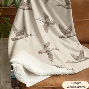 May include: A cream-colored throw blanket with a repeating pattern of brown flying ducks. The blanket features a soft white sherpa fleece lining on the underside. The text "THE" and "S" are partially visible at the bottom of the blanket design.