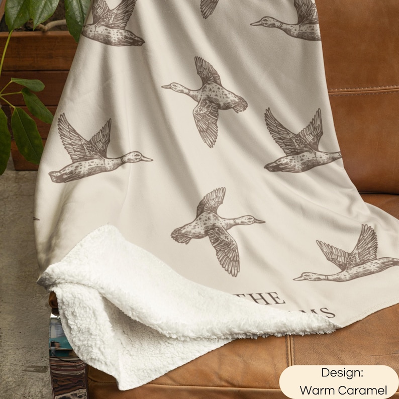 May include: A cream-colored throw blanket with a repeating pattern of brown flying ducks. The blanket features a soft white sherpa fleece lining on the underside. The text "THE" and "S" are partially visible at the bottom of the blanket design.