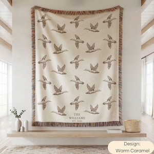 May include: A cream-colored woven throw blanket featuring a repeating pattern of dark brown flying ducks. The bottom center includes the text THE WILLIAMS EST. 2026. The blanket has a multicolored fringed edge on all four sides.