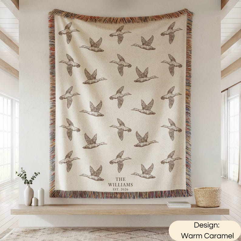 May include: A cream-colored woven throw blanket featuring a repeating pattern of dark brown flying ducks. The bottom center includes the text THE WILLIAMS EST. 2026. The blanket has a multicolored fringed edge on all four sides.