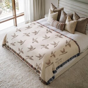 May include: A cream-colored woven throw blanket featuring a repeating pattern of brown flying ducks. The bottom corner includes the text "THE WILLIAMS EST. 2026" in a dark font. The blanket has a multi-colored fringed edge on all sides.