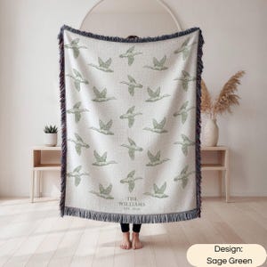 May include: A woven throw blanket featuring a repeating pattern of sage green flying ducks on an off-white background. The blanket has dark blue fringed edges and includes the text THE WILLIAMS EST. 2026 printed in a simple serif font at the bottom.