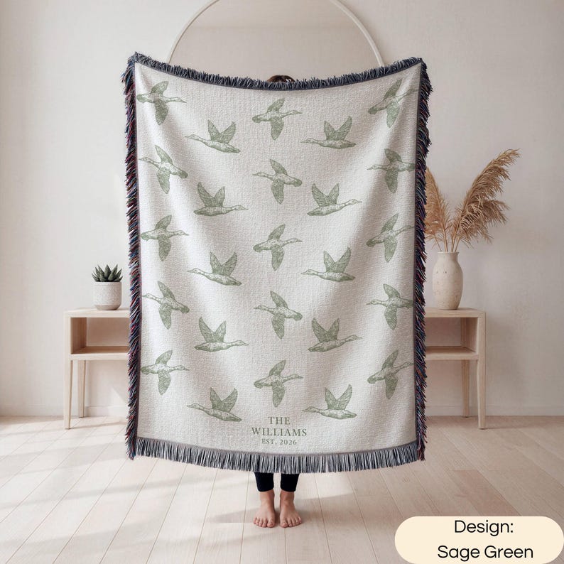 May include: A woven throw blanket featuring a repeating pattern of sage green flying ducks on an off-white background. The blanket has dark blue fringed edges and includes the text THE WILLIAMS EST. 2026 printed in a simple serif font at the bottom.