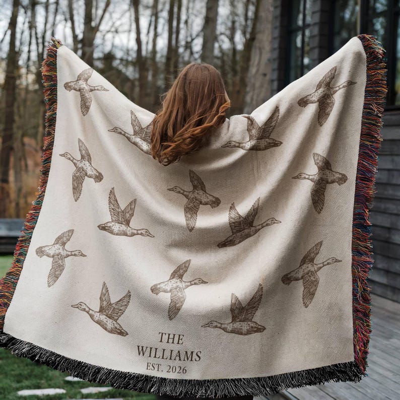 May include: A cream-colored woven throw blanket featuring a repeating pattern of brown flying ducks. The text "THE WILLIAMS EST. 2026" is printed near the bottom. The blanket has a decorative fringe edge with multicolored threads along the sides.