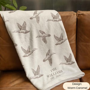 May include: A cream-colored throw blanket featuring a repeating pattern of brown flying ducks. The lower section includes the printed text THE WILLIAMS EST. 2026. The blanket has a soft, plush texture and is draped over a brown leather surface.