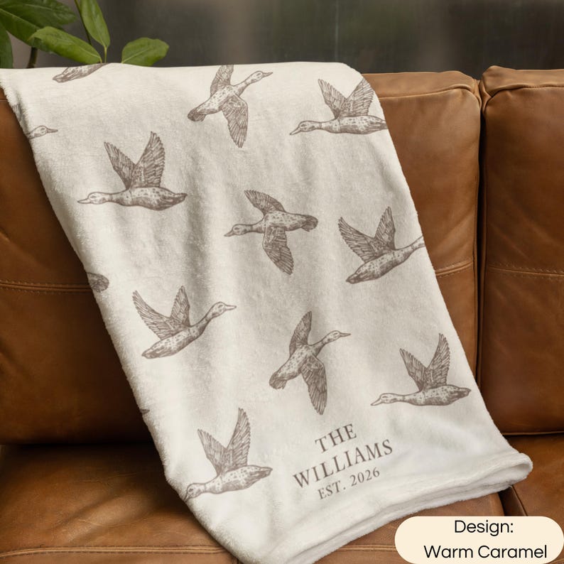 May include: A cream-colored throw blanket featuring a repeating pattern of brown flying ducks. The lower section includes the printed text THE WILLIAMS EST. 2026. The blanket has a soft, plush texture and is draped over a brown leather surface.