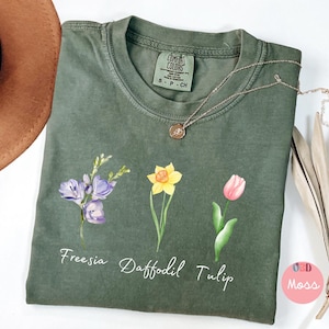 Puede incluir: Una camiseta verde con una ilustración de acuarela de una fresia, un narciso y un tulipán. La camiseta tiene las palabras "Freesia Daffodil Tulip" impresas debajo de las flores.
