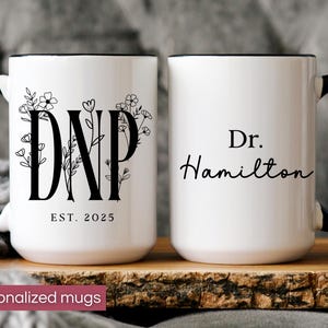 May include: Two white ceramic mugs with black handles and rims. One mug features the letters "DNP" with floral accents and "EST. 2025". The other mug has "Dr. Hamilton" in script. The mugs are on a wooden surface.