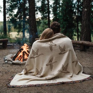 May include: A cream-colored woven throw blanket featuring a repeating pattern of brown flying ducks. The text "THE WILLIAMS EST. 2026" is printed in a dark brown serif font near the bottom edge. The blanket has a fringed edge with multi-colored threads.