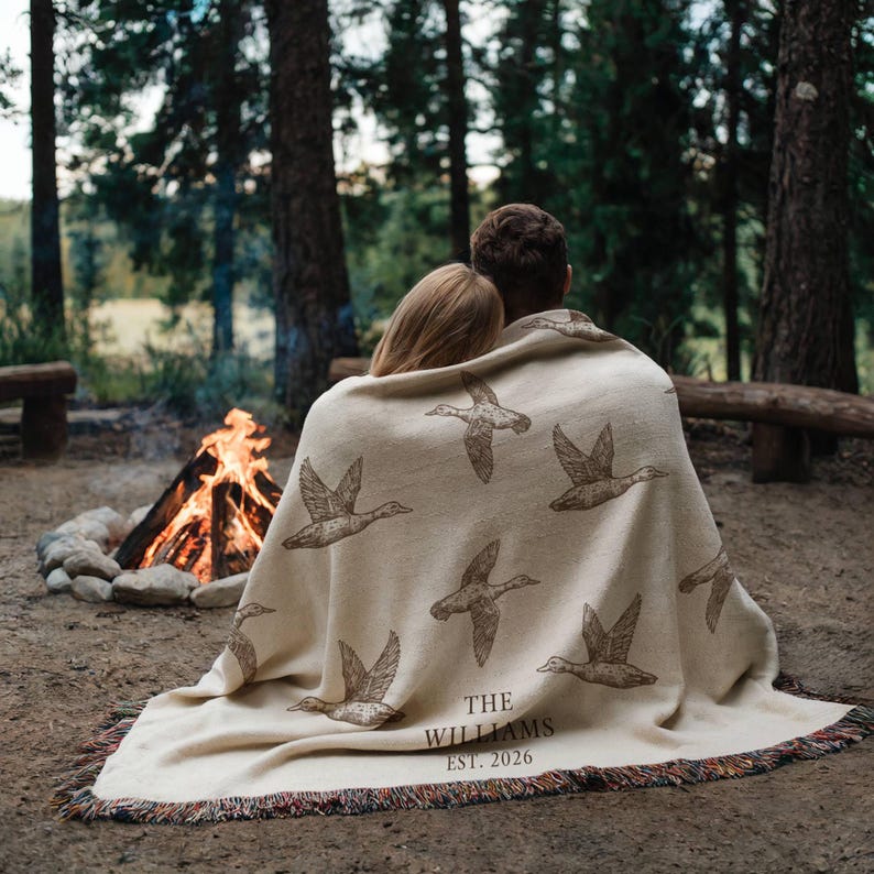 May include: A cream-colored woven throw blanket featuring a repeating pattern of brown flying ducks. The text "THE WILLIAMS EST. 2026" is printed in a dark brown serif font near the bottom edge. The blanket has a fringed edge with multi-colored threads.