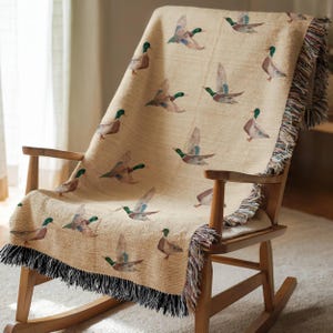 May include: A beige throw blanket with a pattern of flying ducks, draped over a wooden rocking chair. The blanket has a black fringe along the bottom edge and is made of a woven fabric. The ducks are depicted in shades of brown, green, and blue.