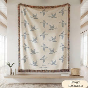 May include: A woven throw blanket with a cream background and a repeating pattern of flying ducks in denim blue. The bottom center features the text THE WILLIAMS EST. 2026. The blanket has a multicolored fringed edge on all four sides.