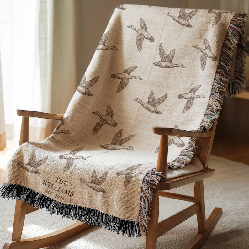 May include: A beige woven throw blanket featuring a repeating pattern of brown flying ducks. The blanket includes the text "THE WILLIAMS EST. 2026" in the bottom left corner and is finished with a multi-colored fringed edge on the sides.